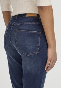 Jean en denim bleu foncé avec une taille mi-haute, présentant un délavage subtil, des coutures claires et deux poches arrière. Étiquette visible au niveau de la taille.