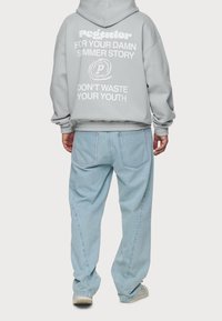 Sweat à capuche gris oversize avec texte blanc au dos, associé à un jean ample bleu clair. Tissu doux, coupe décontractée, poignets et taille élastiqués.