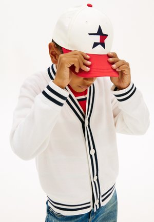 Niño ajustándose una gorra blanca y roja con logo de estrella, vestido con un cárdigan blanco con rayas negras y jeans azules sobre un fondo blanco.