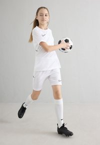 Joven jugadora de fútbol con uniforme blanco, sosteniendo un balón de fútbol y con una pierna levantada, apoyada contra una pared gris lisa.