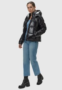 Zwarte pufferjack met een glanzende afwerking, ritssluiting en capuchon. Gedragen over een blauwe top en lichtblauwe jeans, aangevuld met zwarte enkellaarsjes.