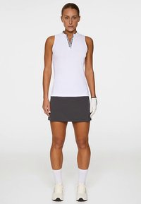 Vit ärmlös topp med delvis dragkedja och logodetalj, matchad med en mörkgrå kjol. Modellen bär vita strumpor och ljusa sportskor.
