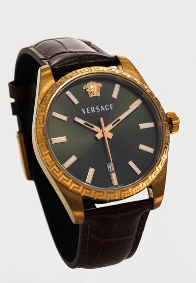Ρολόι χειρός Versace με χρυσή κάσα, σκούρο πράσινο καντράν, χρυσούς δείκτες ώρας, έμβλημα Medusa, παράθυρο ημερομηνίας και καφέ δερμάτινο λουράκι.