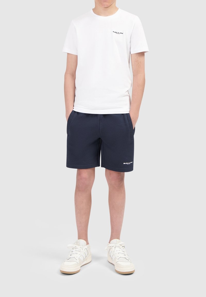 Witte katoenen t-shirt met logo op de borst, gecombineerd met marineblauwe shorts met zakken en een logo. Witte sneakers met accentdetails.