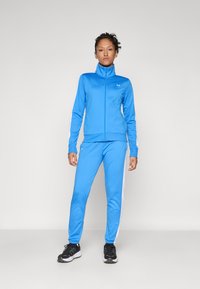 Zils sporta tracksuits ar baltām sānu svītrām; ar rāvējslēdzēju aizdara jaka un atbilstošas bikses no gludas, elastīgas vielas; pieguļošs dizains.