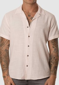 Camisa de manga corta color rosa claro hecha de una tela texturizada, con un cuello y cinco botones marrones en la parte delantera.