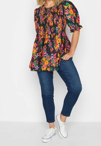 Donna che indossa un top smock floreale colorato con maniche a sbuffo, jeans attillati blu scuro e sneakers bianche stringate, in piedi con una mano in tasca.