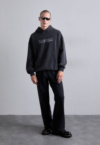 Mann trägt einen schwarzen übergroßen Hoodie mit Text, schwarze weit geschnittene Hose, schwarze Schuhe und dunkle Sonnenbrille vor einer schlichten grauen Wand.