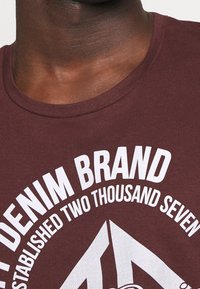 Camiseta de algodón burdeos con cuello redondo; presenta un gran texto gráfico blanco y acentos triangulares en la parte delantera. Textura suave.