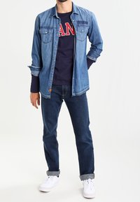Denim-paita, jossa on napitettava etuosa, kaksi rintataskua, käytetty tummansinisellä pitkähihaisella paidalla, jossa on punainen "MAN"-teksti. Tummat farkut ja valkoiset lenkkarit.