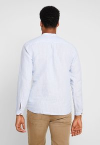 Chemise bleu clair à manches longues avec rayures verticales blanches, dotée d'un empiècement au dos, de poignets à boutons et de fentes latérales. Portée avec un pantalon beige.