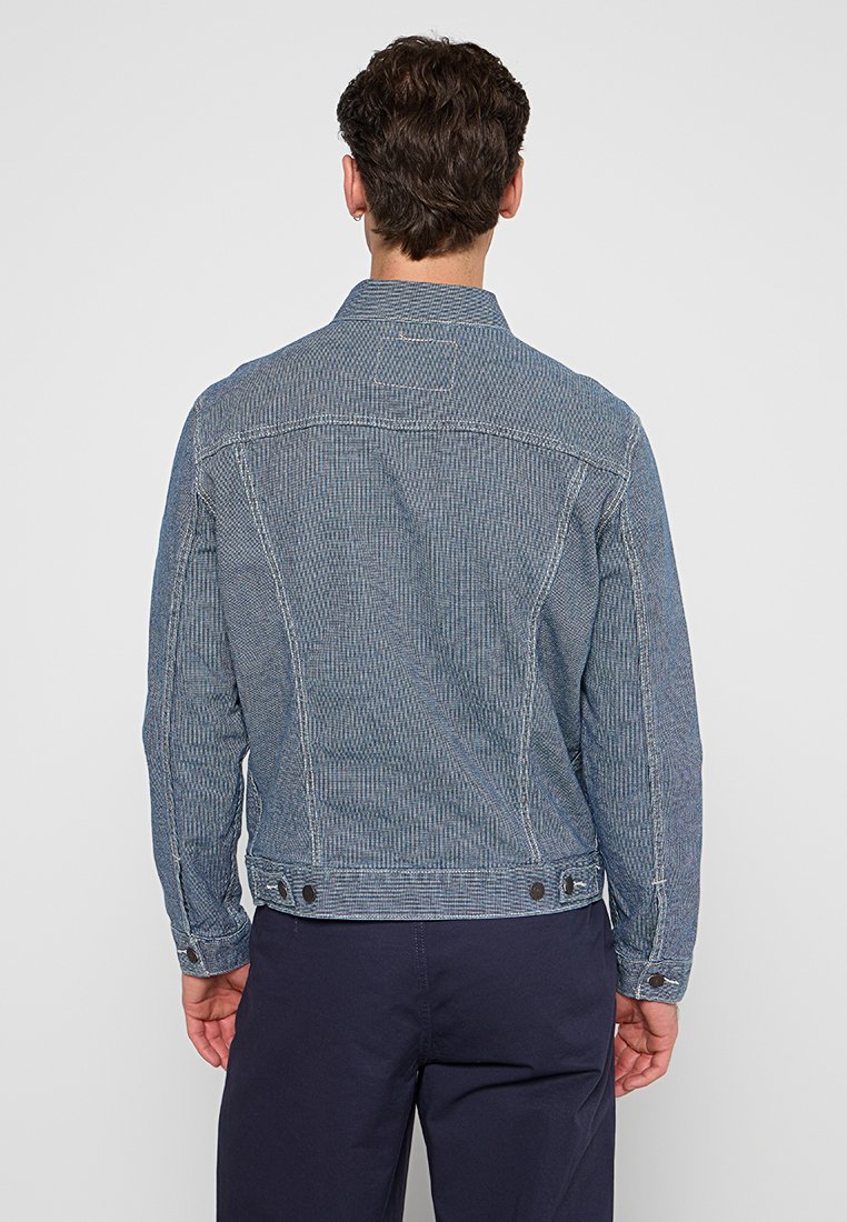 Veste en denim avec un motif chevron, couleur bleu clair et coutures contrastantes. Détails de boutons aux poignets et à l'ourlet.
