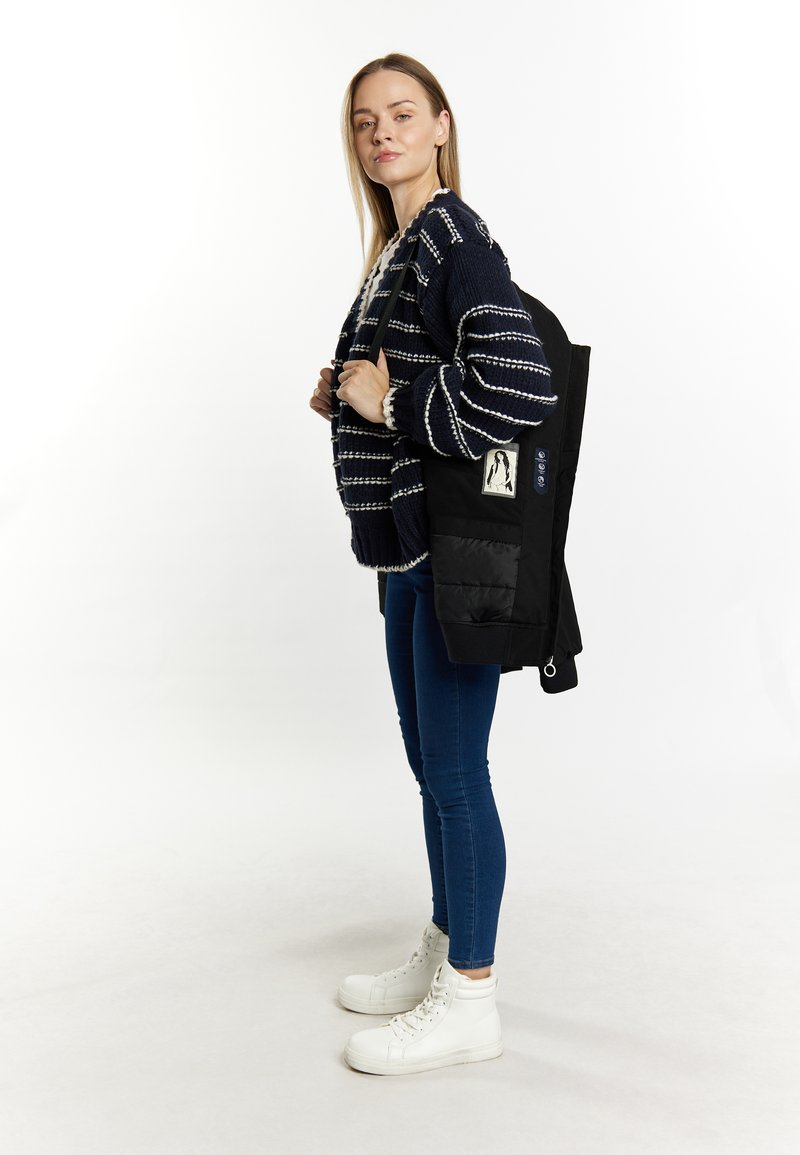 Schwarzer Rucksack mit strukturiertem Design, ausgestattet mit einem Patch und dekorativen Knöpfen, getragen über einem navyblauen gestreiften Pullover mit weißen Akzenten.