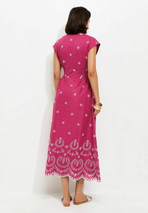 Mujer con un vestido midi sin mangas color magenta con bordados florales blancos, de pie descalza contra un fondo blanco liso.