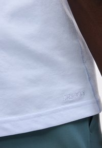 Κοντινή λήψη λευκής ραφής σε πουκάμισο Dri-FIT, φθαρμένο πάνω σε σκούρο πράσινο παντελόνι, με ορατά ράμματα, που δείχνει μέρος του μπράτσου του φορέα.