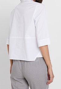 Vit blus med kort ärm och krage, med sömdetaljer över ryggen och rak passform, i kombination med rutiga byxor.