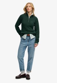 Grüner Strickpullover mit Viertelreißverschluss, hellblauer Streifenhemd darunter, hellblaue Jeans und dunkle burgunderfarbene Mokassins.