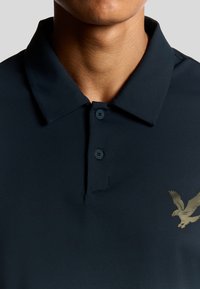 Marinblå polo-shirt med krage, två knappslå och detaljer med en guldörn på vänster bröst. Slät, strukturerad tyg.