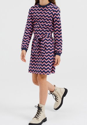 Langärmeliges Kleid mit marineblauem und pinkfarbenem Zickzackmuster, elastischer Taille mit Bindeband, kombiniert mit beigen Schnürstiefeln und schwarzen Sohlen.