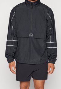 Mężczyzna ubrany w czarną kurtkę typu windbreaker z półzamkiem, ozdobioną białym przeszyciem i logo, w połączeniu z czarnymi szortami, stojący na jednolitym tle.