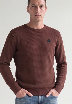 Homme en chemise marron texturée à manches longues, les mains dans les poches, debout devant un fond clair uni.