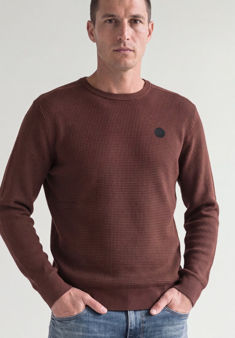 Homme en chemise marron texturée à manches longues, les mains dans les poches, debout devant un fond clair uni.