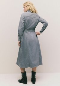 Frau mit lockigem, blonden Haar, die ein graues Denim-Midikleid mit Gürtel und schwarze, genagelte Ankle Boots trägt, steht vor einem schlichten Hintergrund.