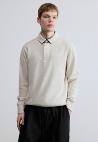 Lys beige langærmet polo med krave, der har knaplukning og tekstureret stof. Kombineret med sorte bukser.