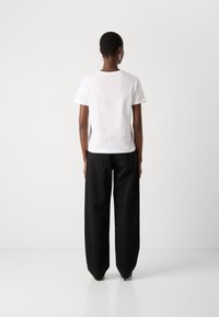 Personne portant un t-shirt blanc uni à manches courtes et un pantalon noir ample, debout de dos devant un fond blanc uni.