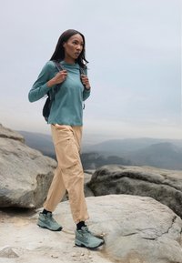 Abbigliamento da escursionismo composto da una maglietta a maniche lunghe color turchese, pantaloni cargo beige e scarponi da trekking grigio-verdi su un terreno roccioso. Include uno zaino nero.