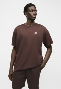 Νέος άνδρας που φοράει καφέ σκούρο t-shirt και σορτς Adidas με λευκά λογότυπα, στέκεται με το ένα χέρι στην τσέπη, κοιτάζοντας δεξιά.