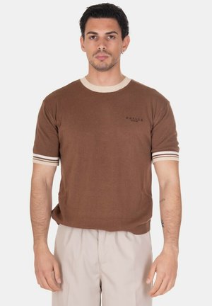 Uomo con capelli corti e scuri indossa una maglia marrone a maniche corte in maglia con rifiniture bianche e pantaloni beige chiaro, in piedi su sfondo bianco.