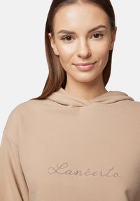 Beżowa bluza z kapturem o miękkiej fakturze z haftowanym napisem "Lanĉerto" w kursywie, mocnymi szwami i luźnym krojem.