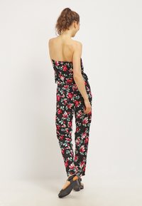 Svart blommig jumpsuit med röda och gröna blommönster, axelbandslös topp, insvängd midja, vida ben och bärs med svarta träskor.