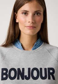 Junge Frau mit langen braunen Haaren, die einen grauen Sweatshirt mit "BONJOUR" in großen dunklen Buchstaben und einem gerafften Jeanskragen darunter trägt.