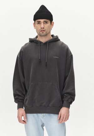 Grauer übergroßer Kapuzenpullover mit einer Fronttasche, Kordelzügen und gerippten Bündchen, kombiniert mit hellblauen Jeans. Eine schwarze Mütze rundet den Look ab.