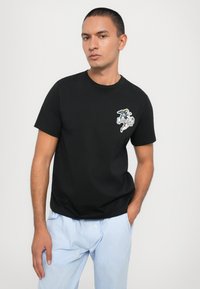 Schwarzes Baumwoll-T-Shirt mit Rundhalsausschnitt und kurzen Ärmeln. Mit einem gestickten Design auf der linken Brust, kombiniert mit hell gestreiften Hosen.