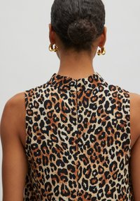 Ärmellose Bluse in Beige mit Leopardenmuster in Braun und Schwarz. Verfügt über einen hohen Kragen und einen Reißverschluss im Rücken, kombiniert mit goldenen Creolen.