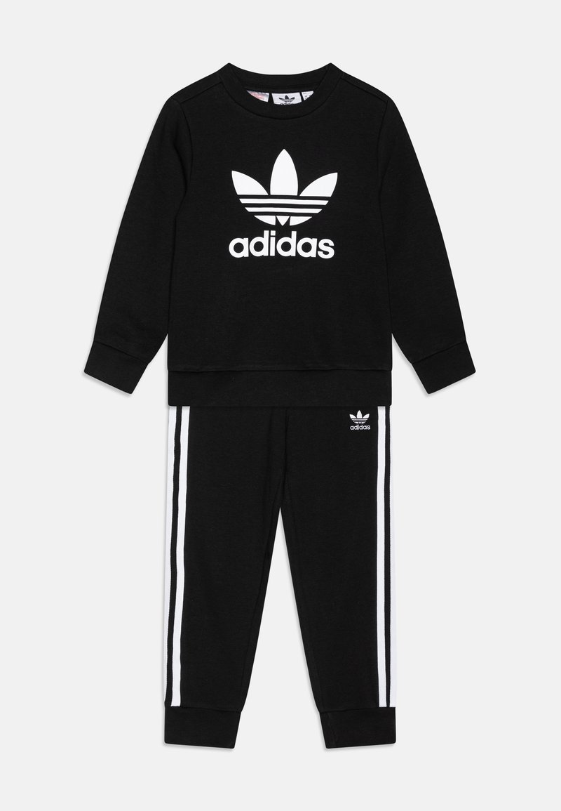 Melnā kokvilnas treniņtērps, kas satur svīteru ar apaļu kaklu un balto Adidas logo, kā arī atbilstošas bikses ar baltām sānu strēmelēm.