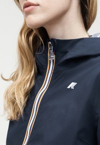 Γυναίκα που φοράει μπλε σκούρο μπουφάν με πολύχρωμο φερμουάρ με ρίγες και λευκό λογότυπο στο στήθος, δείχνοντας το προφίλ της και τα μακριά ξανθά μαλλιά της.