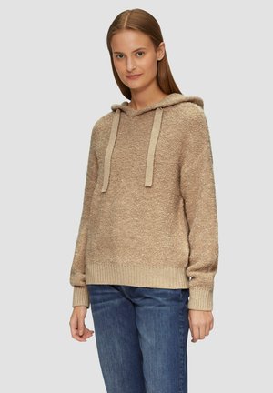 Beige Kapuzenpullover aus strukturiertem Stoff, mit geripptem Saum und Bündchen, ausgestattet mit beigen Kordeln. Kombiniert mit blauen Jeans.