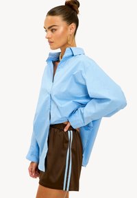 Chemise oversize bleu clair en coton avec un devant boutonné, associée à un short marron avec des rayures bleues sur les côtés.