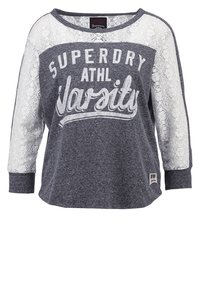 Graues Langarmshirt mit weißen Spitzelementen an den Ärmeln, mit der Aufschrift "SUPERDRY ATHL VARSITY" in fettem weißen Text auf der Vorderseite.