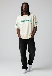 T-shirt beige oversize avec le texte graphique vert "HIRA PARIS" et un design d'étoile sur les manches, associé à un jean noir et des baskets blanches.