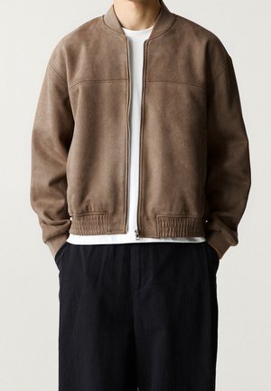 Person iført en brun ruskind bomberjakke med lynlås over en hvid skjorte og sorte bukser, stående med hænderne i lommerne.