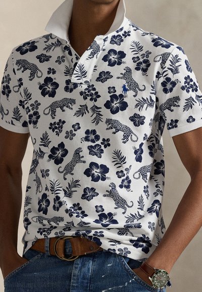 Λευκό πόλο μπλουζάκι με σχέδιο από ναυτικό μπλε λουλουδάτο και λεοπάρ μοτίβο, κοντά μανίκια και αντίθετο γιακά. Φορεμένο με τζιν και καφέ ζώνη.