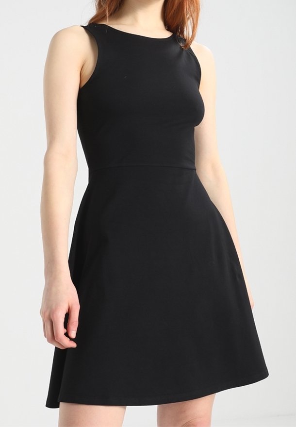 Robe noire sans manches en forme trapèze portée par une femme aux cheveux roux, cintrée à la taille et s'évasant légèrement au niveau de la jupe, sur un fond uni.