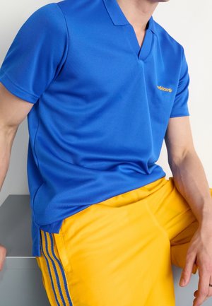 Mann trägt ein blaues Adidas Polohemd und gelbe Hosen mit blauen Streifen, sitzt mit einer Hand auf seinem Bein.