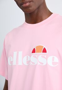 Rozā T-krekls ar baltu "ellesse" logo un teksturētu, daudzkrāsainu logo detaļu sarkanā, oranžā un baltā krāsā. Ikdienas piegriezums un īsas piedurknes.