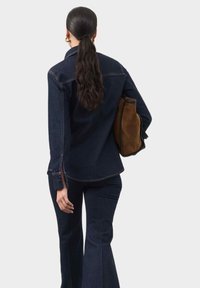 Mørk denimjakke med let flæse i kanten, parret med matchende flared bukser. Holder en brun ruskinds taske. Glat tekstur, minimal syning.